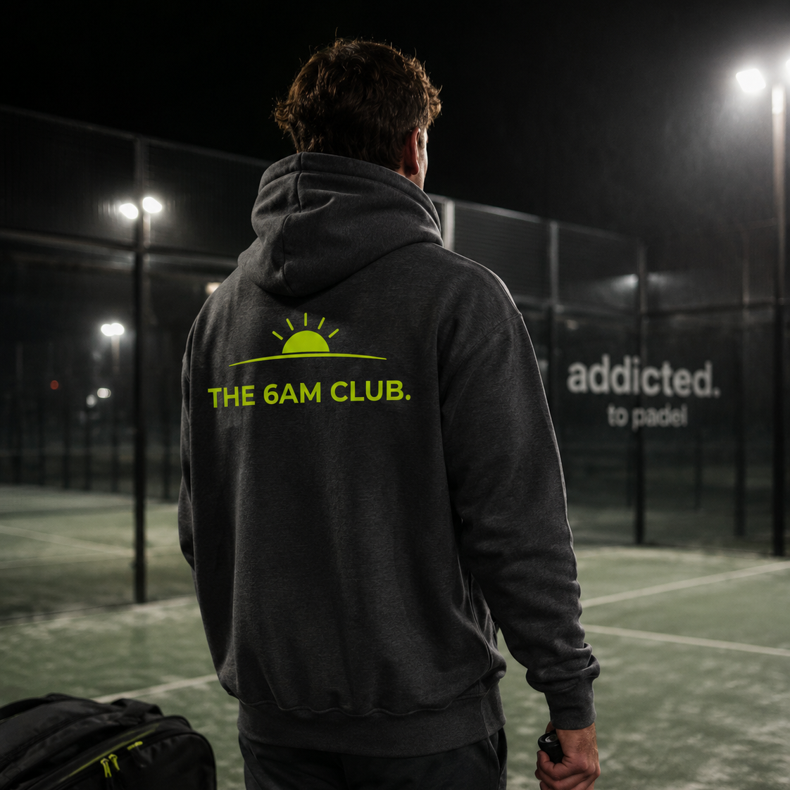 Person wearing a hoodie with 'The 6AM Club' logo on a padel court at night.