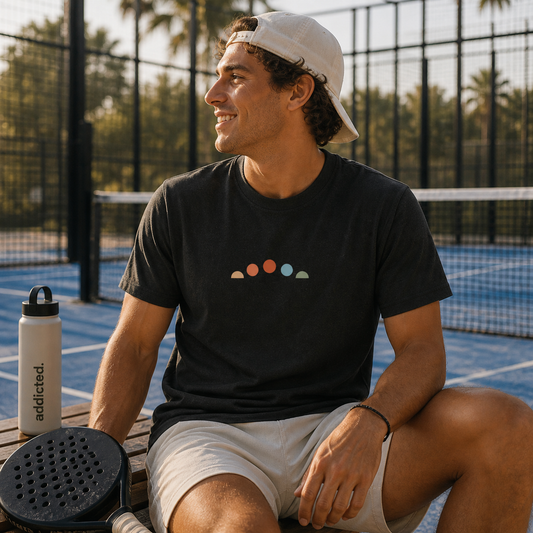 Man sitting on a tennis court with a water bottle and racket, wearing a black t-shirt with colorful design and a white cap.