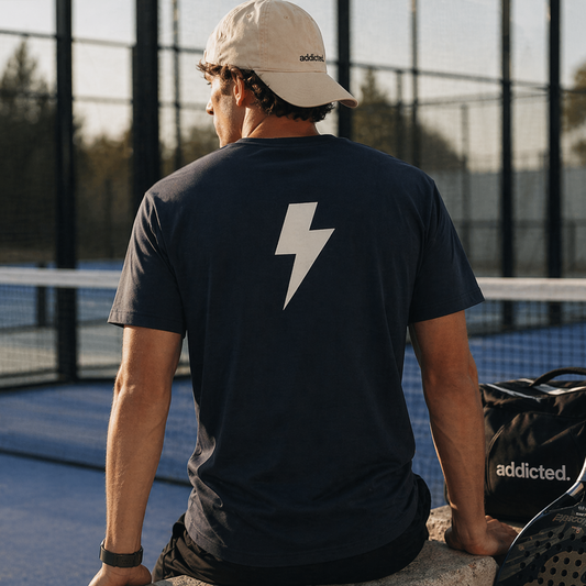 Person sitting on a tennis court wearing a black t-shirt with a white lightning bolt design, holding a racket.