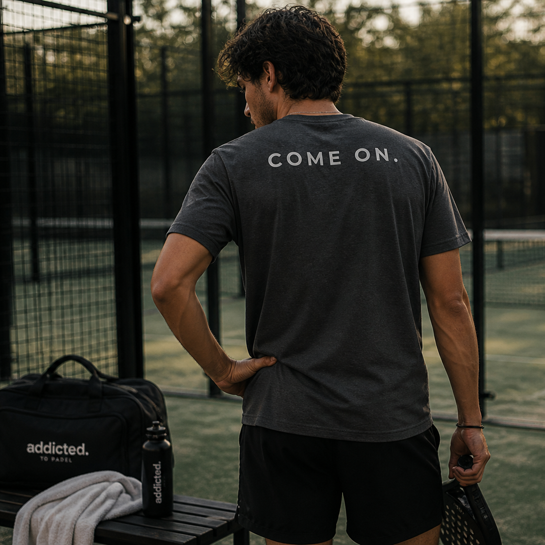 Man standing on a padel court wearing a black t-shirt with 'COME ON.' text, holding a racket.