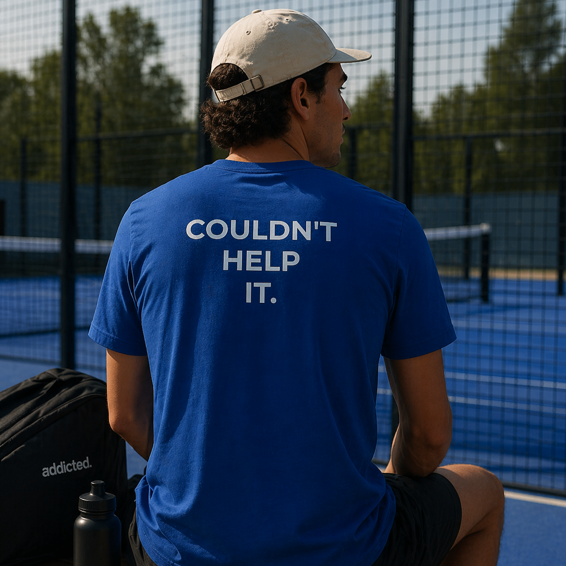 Person wearing a blue t-shirt with 'COULDN'T HELP IT.' text, sitting on a bench at a sports court.