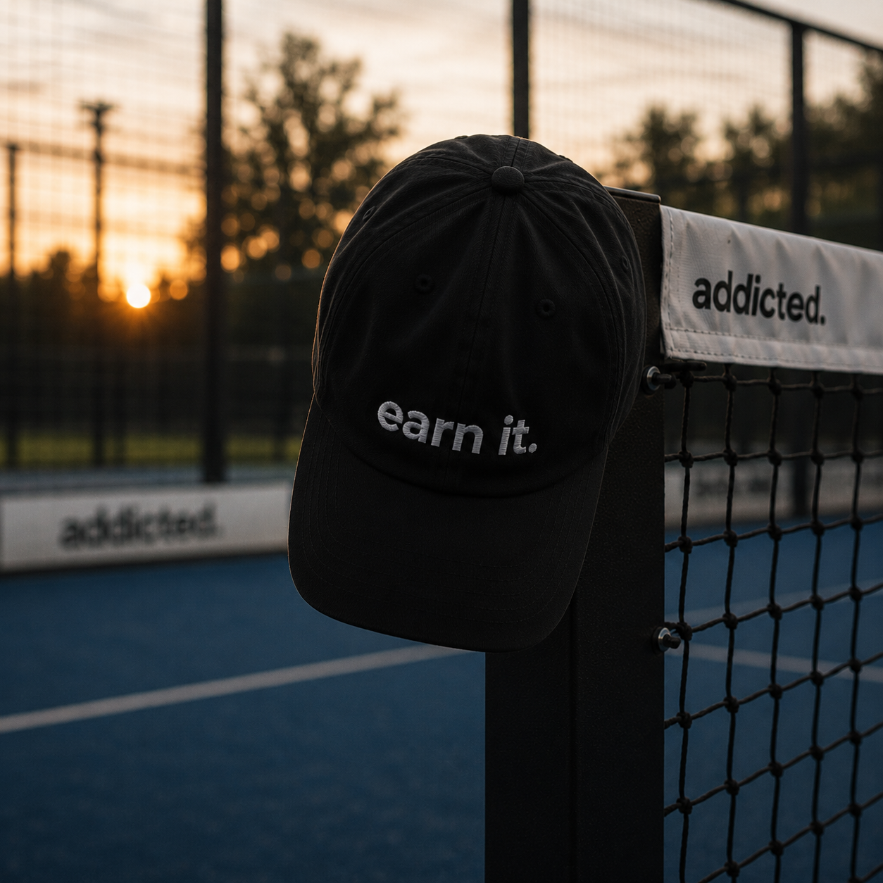 Black cap with 'earn it.' text on a tennis court at sunset
