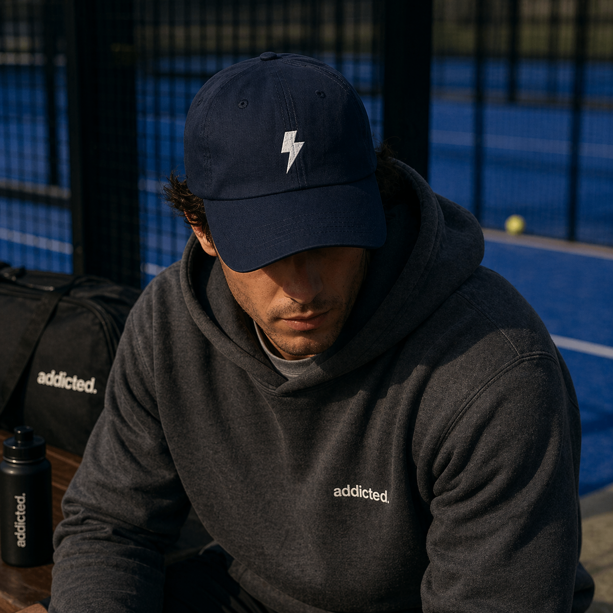 Man wearing a navy cap with a white logo and gray 'addicted' hoodie on a blue court.