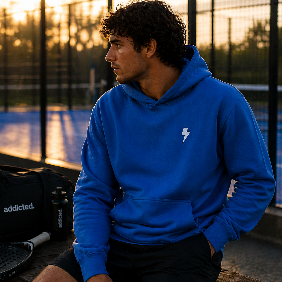 Person wearing a blue hoodie with a lightning bolt logo on a sports court at sunset.