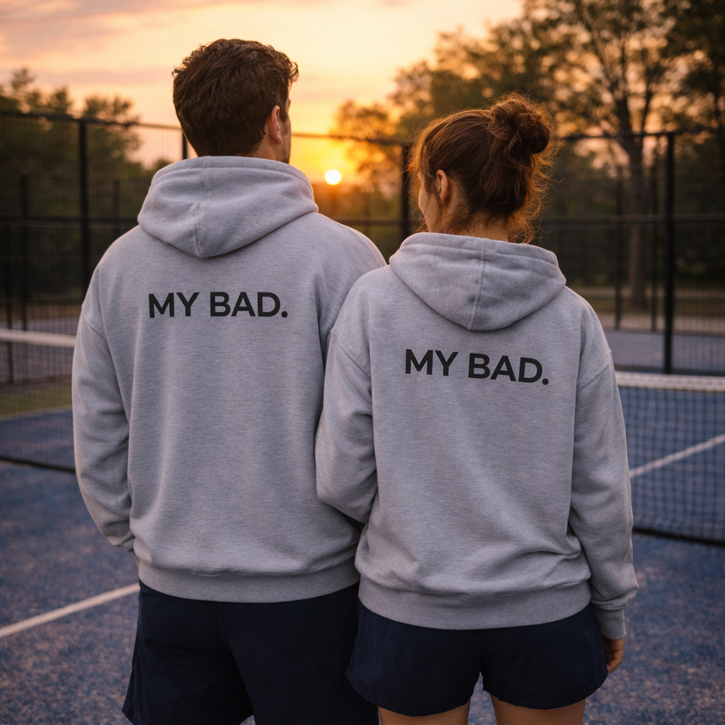 Two people wearing gray hoodies with 'MY BAD.' text on a tennis court at sunset.