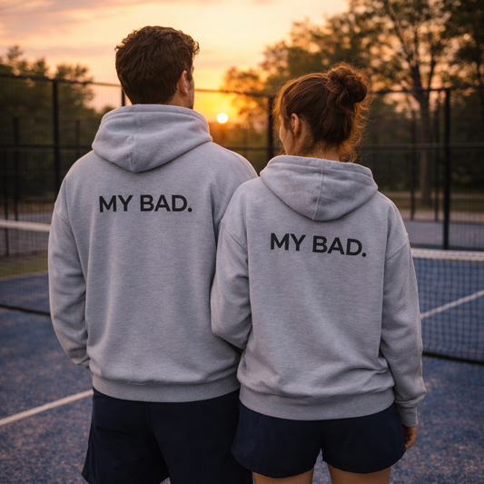 Two people wearing gray hoodies with 'MY BAD.' text on a tennis court at sunset.