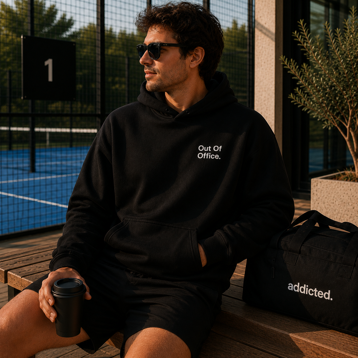 A man wearing a black hoodie with the white text 'Out Of Office.' sits on a wooden bench in front of a tennis court.