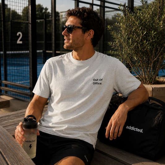 A man wearing a light gray t-shirt with the text 'Out Of Office.' on the chest, sitting on a wooden bench next to a black 'addicted.
