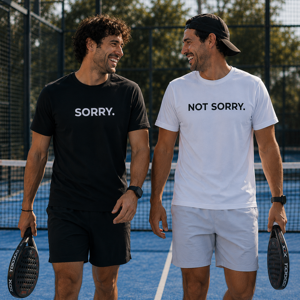 Two men on a tennis court, one wearing a black 'SORRY.' shirt and the other in a white 'NOT SORRY.' shirt.