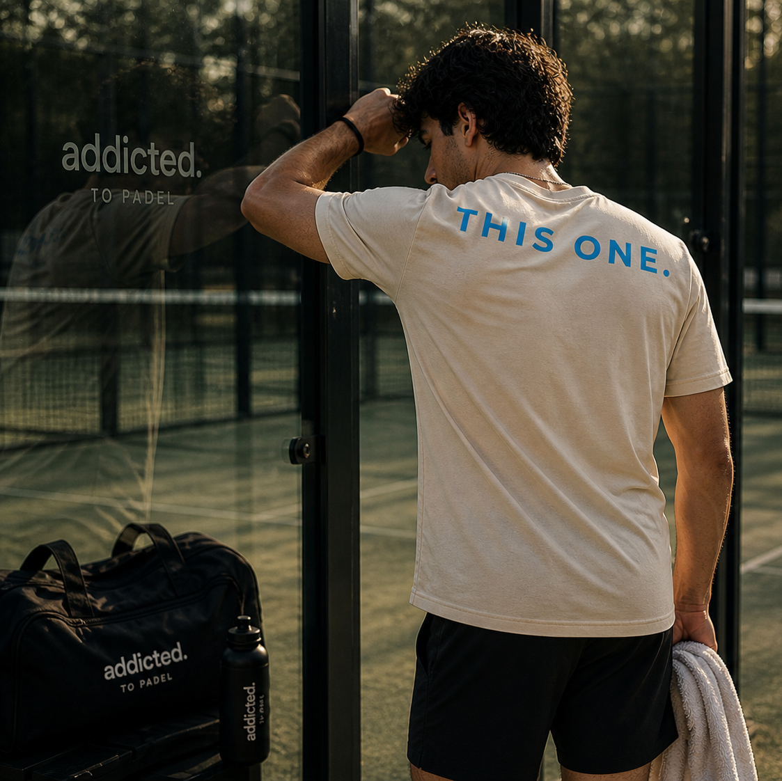 Man on a padel court wearing a shirt with 'This One' text, standing next to an 'addicted. To Padel' bag.
