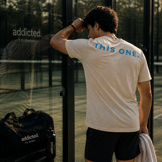 Man on a padel court wearing a shirt with 'This One' text, standing next to an 'addicted. To Padel' bag.