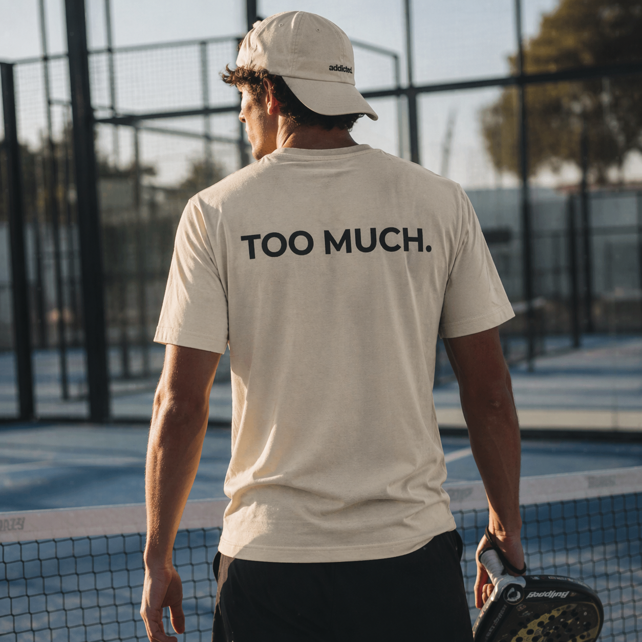 Man on a tennis court wearing a beige t-shirt with 'Too Much.' text.