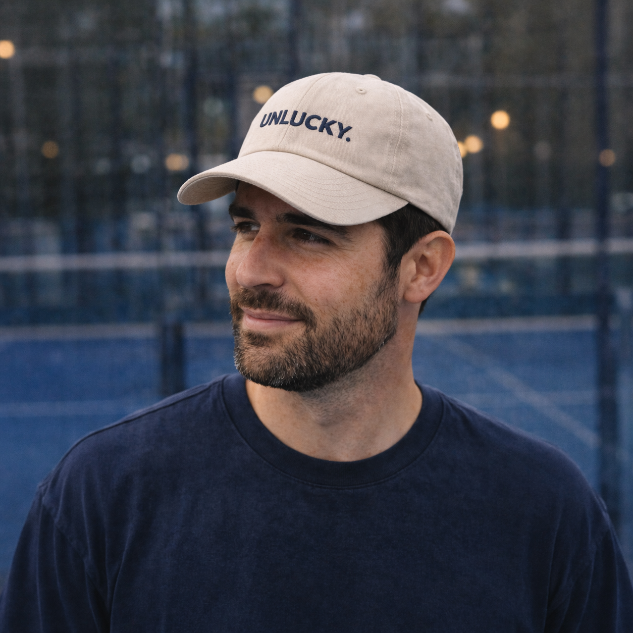 Man wearing a beige cap with 'UNLUCKY.' text on a tennis court.