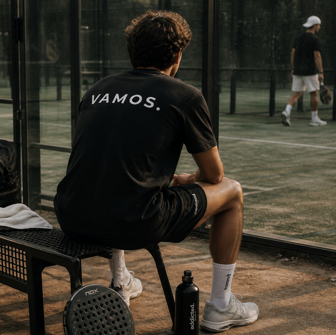 Person sitting on a bench with 'VAMOS.' shirt at a tennis court