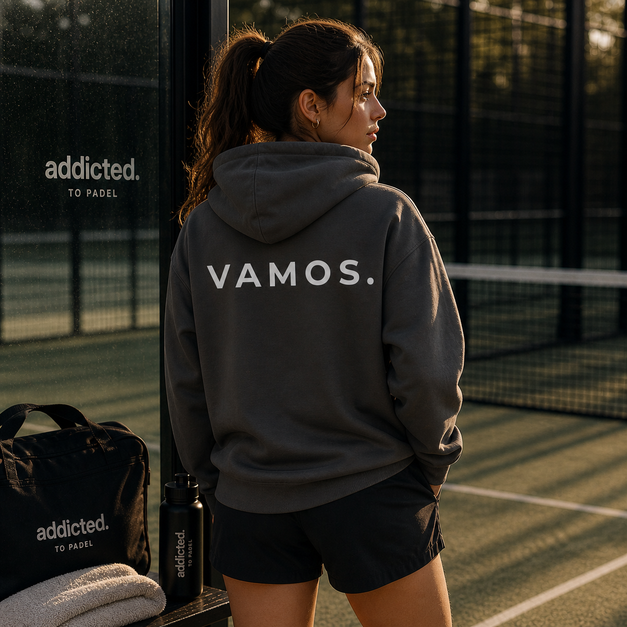 Person wearing a hoodie with 'VAMOS.' on a padel court, with 'addicted.' branding visible.