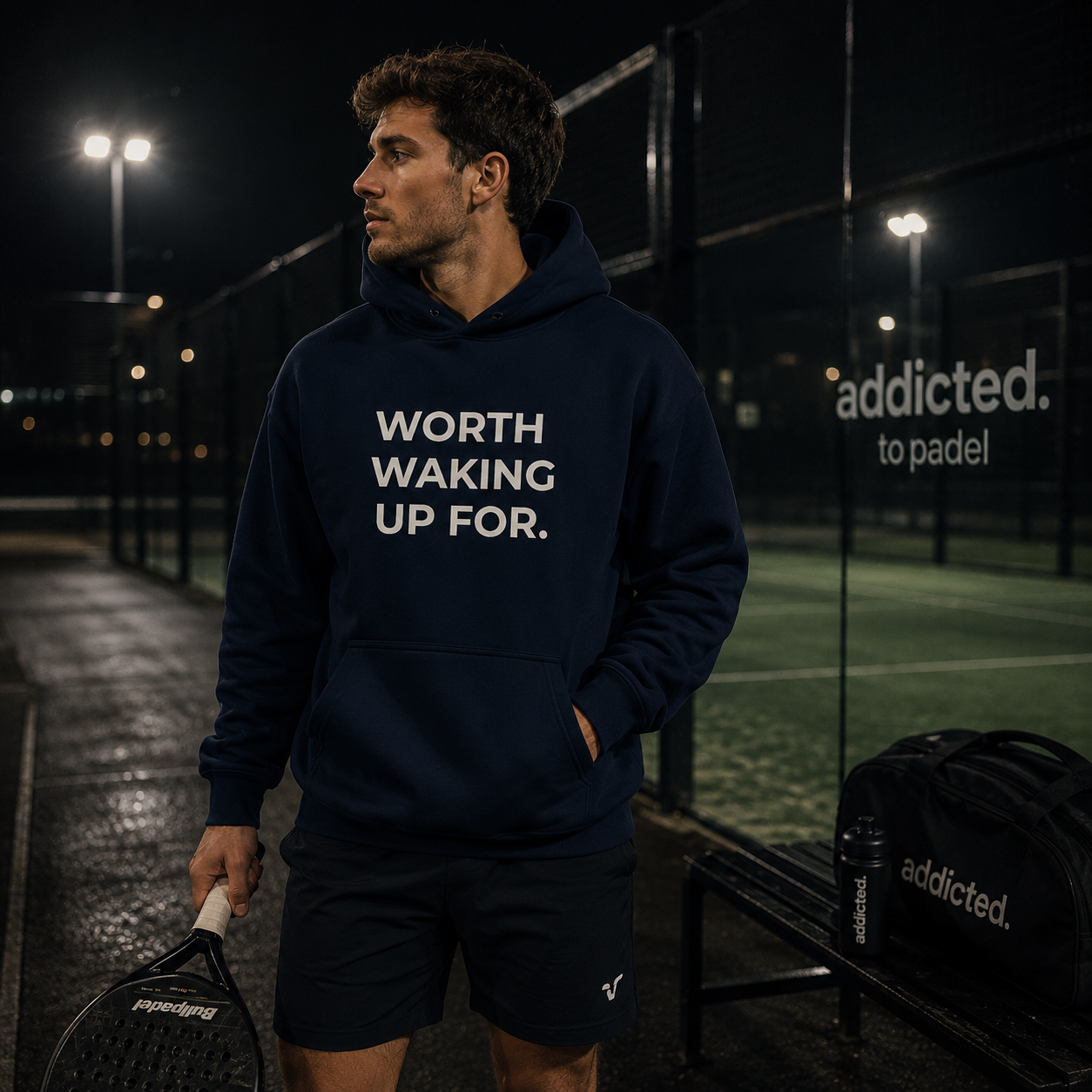 Man wearing a navy hoodie with 'WORTH WAKING UP FOR.' text on a padel court at night.