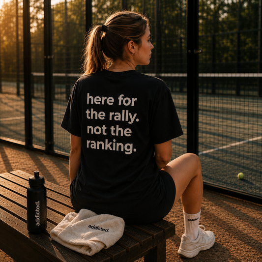 Person sitting on a bench with a t-shirt reading 'here for the rally. not the ranking.' on a tennis court.
