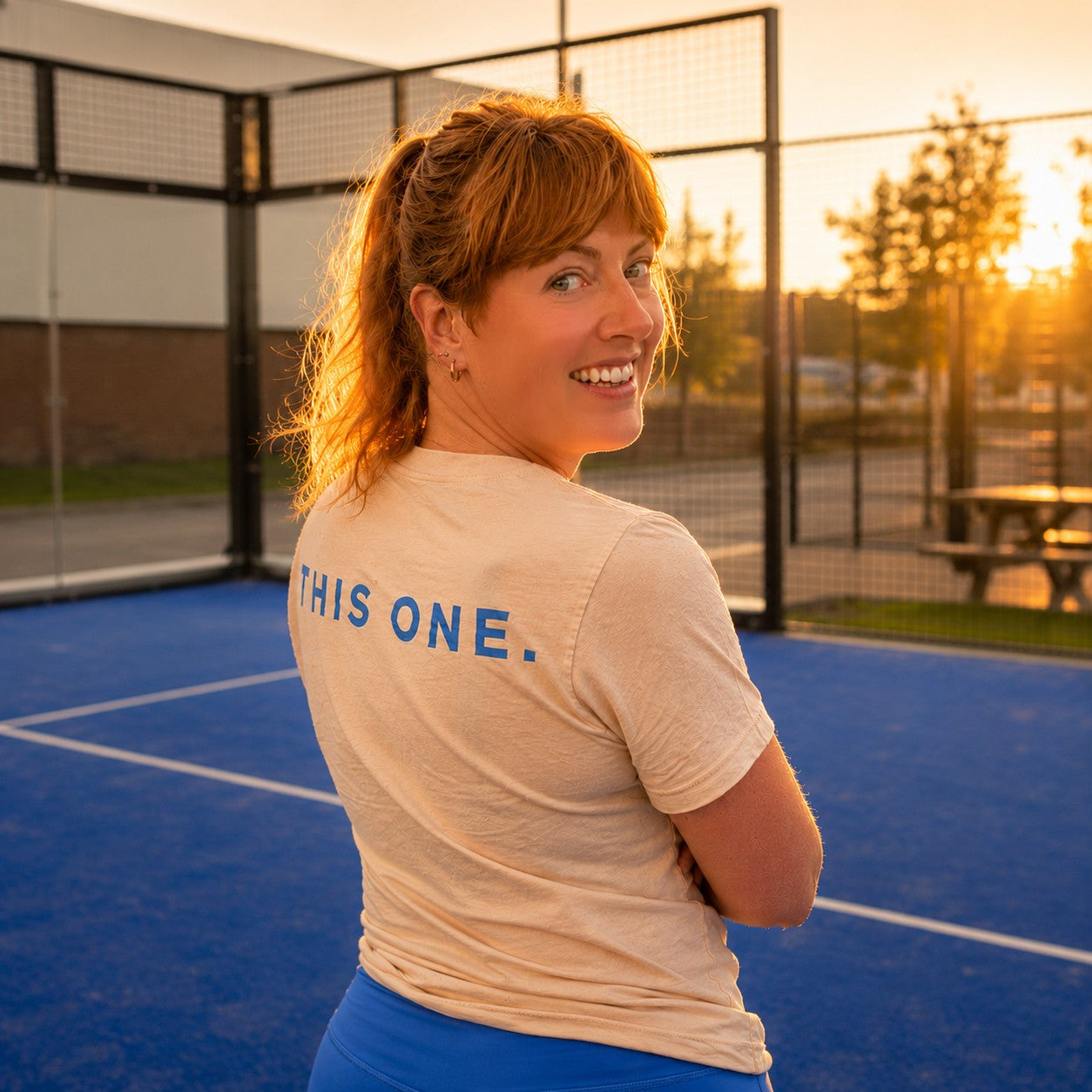Woman standing on a blue tennis court with 'THIS ONE.' shirt, sunset in background