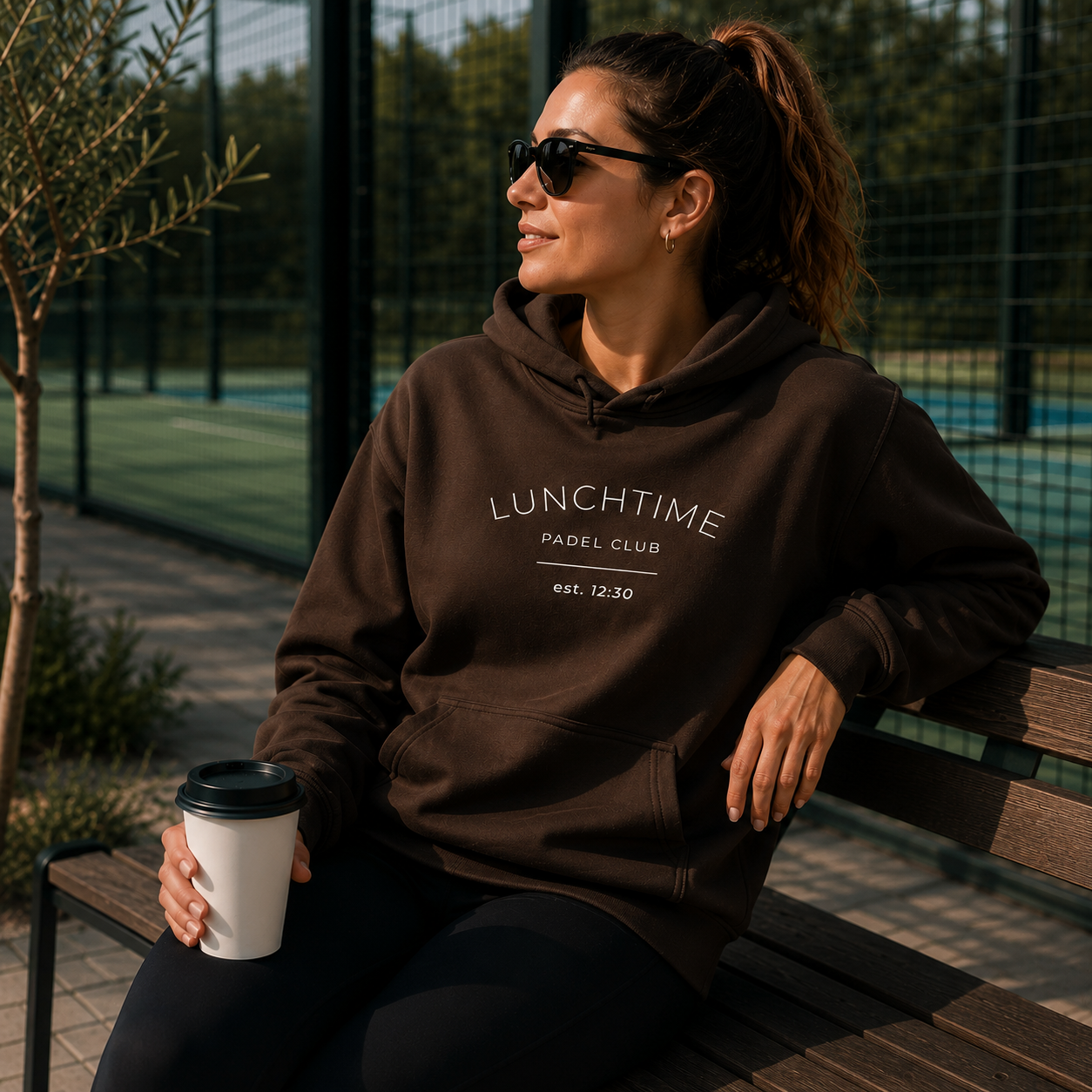 A woman wearing a dark brown pullover hoodie with 'LUNCHTIME PADEL CLUB est. 12:30' printed in white on the chest, sitting on a wooden bench at an outdoor tennis court.