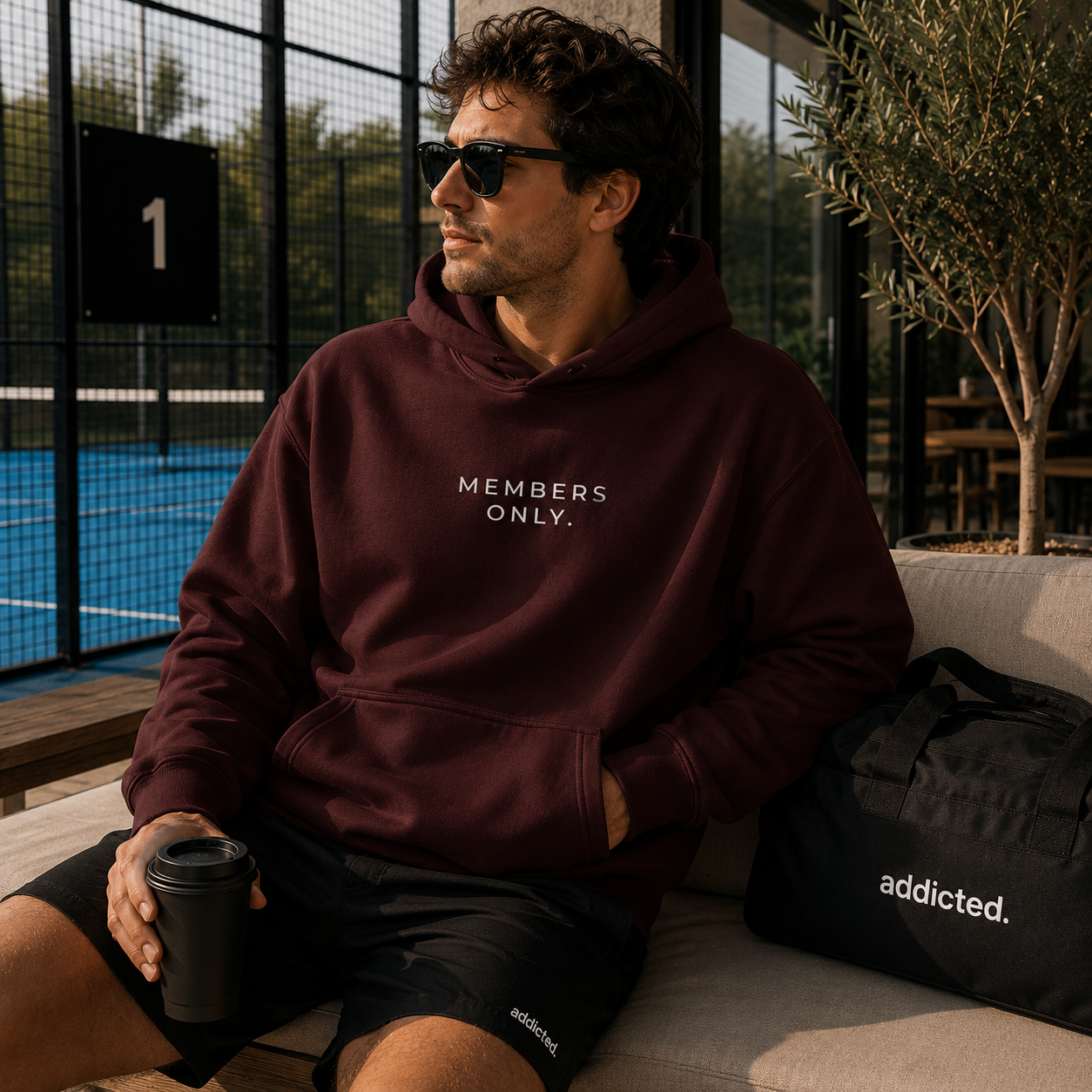 A man wearing a burgundy hoodie with 'MEMBERS ONLY.' printed in white on the chest sits on a beige outdoor sofa.