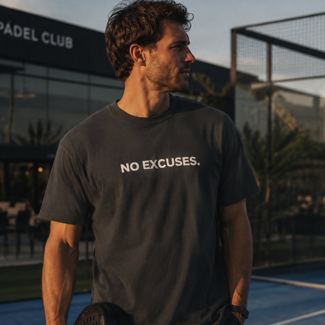A man on a blue padel court wearing a dark charcoal gray t-shirt with the white text 'NO EXCUSES.' printed across the chest, holding a padel racket.