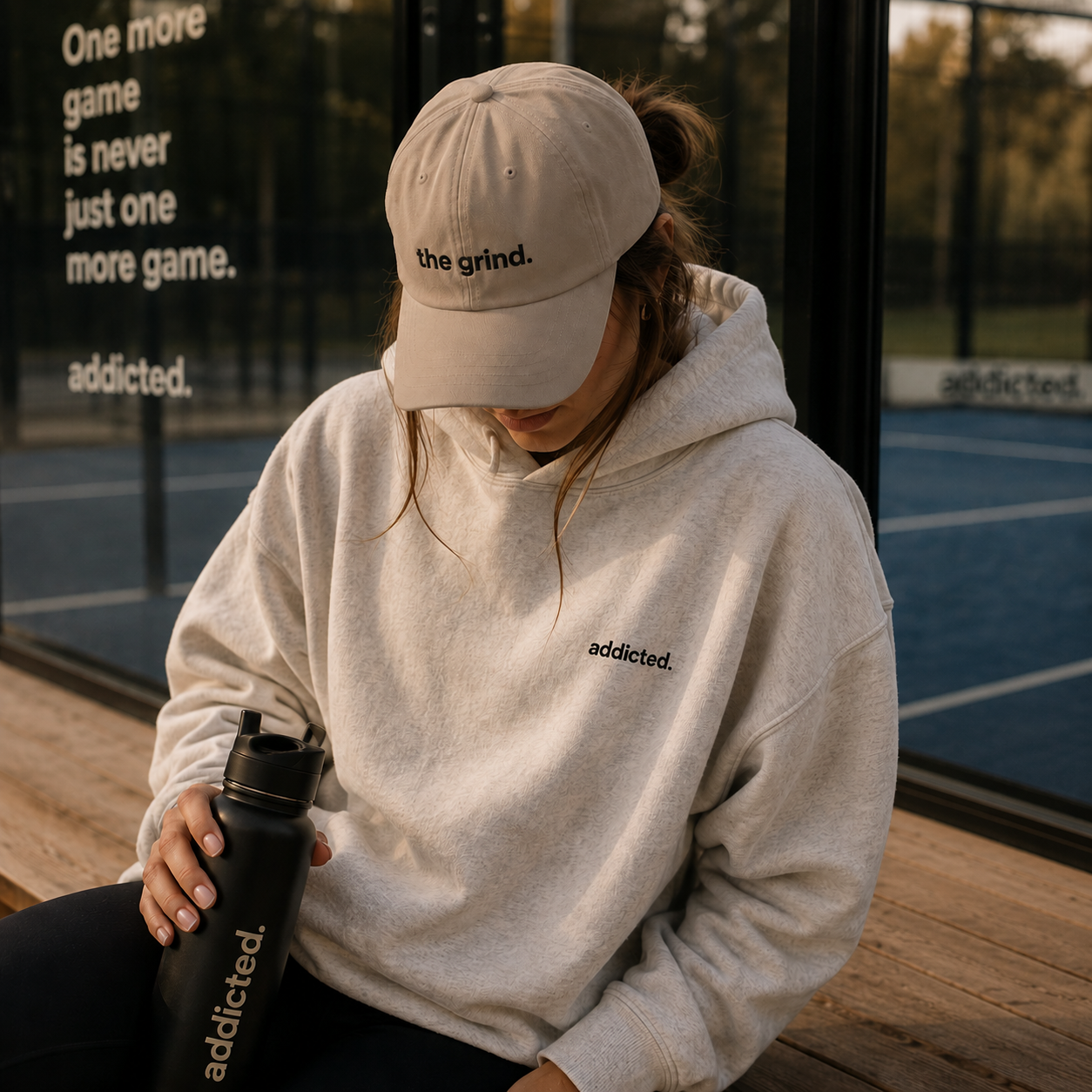 Person wearing a beige cap and hoodie with 'addicted' branding, sitting on a wooden bench.